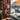 A cup of latte art coffee sits on a small blue saucer on a wooden windowsill. Next to the coffee is a decorative vase filled with autumn flowers and several small pumpkins. Outside the window is a blurred street scene showing buildings and trees with colorful fall foliage.