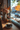 A cup of latte art coffee sits on a small blue saucer on a wooden windowsill. Next to the coffee is a decorative vase filled with autumn flowers and several small pumpkins. Outside the window is a blurred street scene showing buildings and trees with colorful fall foliage.