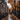A medium shot depicts two people walking away from the viewer on a wet, brick-paved path lined with orange leaves. The path is flanked by elegant, brick buildings on the left and trees with bright orange leaves on the right. The wet path reflects the buildings, trees and sky above.