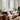 A bright and stylish living room centered around a large brown leather sofa piled with decorative pillows and a light-colored throw blanket. Two tall, arched windows dominate the background, allowing natural light to flood the room and reveal green trees outside. Wooden side and coffee tables, potted plants, a floor lamp, and a patterned rug complete this inviting and well-decorated space.