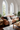 A bright and stylish living room centered around a large brown leather sofa piled with decorative pillows and a light-colored throw blanket. Two tall, arched windows dominate the background, allowing natural light to flood the room and reveal green trees outside. Wooden side and coffee tables, potted plants, a floor lamp, and a patterned rug complete this inviting and well-decorated space.