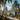 A serene view of a tropical resort featuring a row of modern, white rectangular bungalows with large glass windows, nestled among tall palm trees and various lush green foliage. A light-colored gravel path winds through the foreground, flanked by rocks and plants, leading towards distant green, forested hills under a bright blue sky with fluffy white clouds, suggesting a peaceful, luxurious vacation spot.