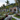 A picturesque view of modern white bungalows with dark roofs and large glass windows, nestled in a vibrant tropical garden. The garden features colorful flowers, green plants, and large rocks, with a light gravel path winding between them. Tall palm trees frame the scene, with a dense, green jungle-covered hill in the background under a softly lit sky, creating a peaceful and luxurious resort atmosphere.