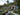 A picturesque view of modern white bungalows with dark roofs and large glass windows, nestled in a vibrant tropical garden. The garden features colorful flowers, green plants, and large rocks, with a light gravel path winding between them. Tall palm trees frame the scene, with a dense, green jungle-covered hill in the background under a softly lit sky, creating a peaceful and luxurious resort atmosphere.