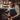 A vertical, medium shot depicts a male barista with thick, curly brown hair, a full beard, and black-framed glasses. He is wearing a light-colored striped shirt and a dark apron, intently making coffee using a black espresso machine. His left hand is on the machine's lever, and his right hand holds a white cup with a coffee beverage, featuring subtle latte art, on the machine's drip tray. The background is blurred, showing a warm-toned cafe interior with wooden shelving.