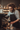 A vertical, medium shot depicts a male barista with thick, curly brown hair, a full beard, and black-framed glasses. He is wearing a light-colored striped shirt and a dark apron, intently making coffee using a black espresso machine. His left hand is on the machine's lever, and his right hand holds a white cup with a coffee beverage, featuring subtle latte art, on the machine's drip tray. The background is blurred, showing a warm-toned cafe interior with wooden shelving.