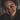 A close-up, slightly angled portrait of a young woman with a genuine, radiant smile revealing white teeth. Her face is richly covered in freckles, with rosy cheeks, and bright blue-green eyes that sparkle. Her reddish-brown hair softly frames her face, and she wears a dark, possibly knit, sweater, all captured in soft, natural lighting against a softly blurred background.