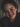 A close-up, slightly angled portrait of a young woman with a genuine, radiant smile revealing white teeth. Her face is richly covered in freckles, with rosy cheeks, and bright blue-green eyes that sparkle. Her reddish-brown hair softly frames her face, and she wears a dark, possibly knit, sweater, all captured in soft, natural lighting against a softly blurred background.