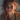 A close-up portrait of a young woman with striking wavy red hair, light freckles, and warm brown eyes, looking directly at the viewer with a genuine, gentle smile. Soft natural light illuminates her face from the left, highlighting her features and creating a bright, inviting expression against a blurred, neutral background.
