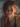A close-up portrait of a young woman with striking wavy red hair, light freckles, and warm brown eyes, looking directly at the viewer with a genuine, gentle smile. Soft natural light illuminates her face from the left, highlighting her features and creating a bright, inviting expression against a blurred, neutral background.