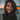 A close-up portrait of a young woman with long, dark, windblown hair smiling genuinely at the viewer. Her face is illuminated by warm, natural light, highlighting her wide smile and faint freckles across her nose and cheeks. The background is a soft, out-of-focus blend of muted blue-gray and warm orange bokeh lights, suggesting a serene sunset or dusk setting.