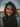 A close-up portrait of a young woman with long, dark, windblown hair smiling genuinely at the viewer. Her face is illuminated by warm, natural light, highlighting her wide smile and faint freckles across her nose and cheeks. The background is a soft, out-of-focus blend of muted blue-gray and warm orange bokeh lights, suggesting a serene sunset or dusk setting.