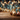 A close-up photograph of festive holiday decorations arranged on a rustic wooden surface. In the lower right, a glittery blue gift box with a gold ribbon, a gold glitter ball, a rose gold glitter ball, and two brown pinecones are scattered among gold confetti. The background is a soft blur of warm gold and cool blue bokeh lights, speckled with subtle star-shaped sparkles.
