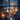 A vertical shot through a slightly wet window pane, looking into a cozy indoor Christmas scene at dusk or night. Inside, a brightly lit Christmas tree adorned with warm white lights and white ornaments stands tall, with wrapped gifts at its base, while a mother and child sit on a rug beside it, illuminated by the warm glow. Outside the window, blurred bare trees are visible against a dark blue sky, and glowing orange bokeh lights shimmer in the foreground, creating a magical and intimate holiday atmosphere.