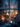 A vertical shot through a slightly wet window pane, looking into a cozy indoor Christmas scene at dusk or night. Inside, a brightly lit Christmas tree adorned with warm white lights and white ornaments stands tall, with wrapped gifts at its base, while a mother and child sit on a rug beside it, illuminated by the warm glow. Outside the window, blurred bare trees are visible against a dark blue sky, and glowing orange bokeh lights shimmer in the foreground, creating a magical and intimate holiday atmosphere.