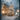 A vertical image depicting a snow-covered Christmas tree outside a rustic building on a magical winter night. The scene is viewed through a window, with blurred ice and snow particles creating a sparkling bokeh effect in the dark foreground. The evergreen tree is brightly lit with warm string lights and silver ornaments, with wrapped gifts nestled at its snowy base, while a warm glow emanates from a large window of the adjacent building, also filled with festive lights and decorations.
