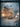 A vertical image depicting a snow-covered Christmas tree outside a rustic building on a magical winter night. The scene is viewed through a window, with blurred ice and snow particles creating a sparkling bokeh effect in the dark foreground. The evergreen tree is brightly lit with warm string lights and silver ornaments, with wrapped gifts nestled at its snowy base, while a warm glow emanates from a large window of the adjacent building, also filled with festive lights and decorations.