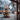 A vertical photograph looking out through a frosted window pane during winter. Indoor evergreen branches decorated with warm white Christmas lights and a red ornament are visible in the foreground. Outside, an out-of-focus snow-covered yard features a large Christmas tree illuminated with golden lights, with a distant house also glowing faintly in the background.