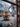 A vertical photograph looking out through a frosted window pane during winter. Indoor evergreen branches decorated with warm white Christmas lights and a red ornament are visible in the foreground. Outside, an out-of-focus snow-covered yard features a large Christmas tree illuminated with golden lights, with a distant house also glowing faintly in the background.