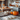 A warm kitchen scene showcasing the preparation of baked goods for the fall season. On a wooden countertop is a cutting board with prepared dough dusted with flour, next to a rolling pin and three pumpkins: a large orange one, a small one, and a white pumpkin. The kitchen is out of focus in the background, with pumpkins and flowers visible on the countertops and shelves.