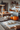 A warm kitchen scene showcasing the preparation of baked goods for the fall season. On a wooden countertop is a cutting board with prepared dough dusted with flour, next to a rolling pin and three pumpkins: a large orange one, a small one, and a white pumpkin. The kitchen is out of focus in the background, with pumpkins and flowers visible on the countertops and shelves.