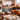 A close-up of uncooked dough dusted with flour and a wooden rolling pin, set on a wooden countertop. In the blurred background, several pumpkins of varying sizes and colors rest on the counter of a bright kitchen. Cabinets and jars are also visible in the background.