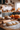 A close-up of uncooked dough dusted with flour and a wooden rolling pin, set on a wooden countertop. In the blurred background, several pumpkins of varying sizes and colors rest on the counter of a bright kitchen. Cabinets and jars are also visible in the background.