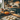 A medium shot of a wooden table in a cozy kitchen, on which six balls of dough covered in flour sit arranged in rows.  A wooden rolling pin lies to the right of the dough, and decorative orange and white pumpkins are arranged on a wooden cutting board in the background. The backdrop is a blurred kitchen with black cabinets and wooden shelves.