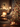 A warm, inviting indoor scene of a Christmas living room at night. A decorated Christmas tree with red baubles and glowing lights stands prominently on the right, with several lit pillar candles and wrapped gifts at its base on a wooden floor. In the softly blurred background, a fireplace with a cozy fire and a lighted garland, along with more lit candles on a coffee table, complete the festive holiday ambiance.