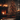 A warmly lit indoor scene at night, featuring a tall, dark green Christmas tree heavily decorated with numerous small, bright lights and shiny red ball ornaments. To the left, a brick fireplace has a roaring, glowing fire, and its mantelpiece is adorned with several lit candles, green garland, and red stockings. Wrapped gifts sit beneath the tree on a wooden floor, next to a light-colored textured rug, while blurred candles in the foreground add to the soft, intimate glow of the setting.