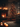 A warmly lit indoor scene at night, featuring a tall, dark green Christmas tree heavily decorated with numerous small, bright lights and shiny red ball ornaments. To the left, a brick fireplace has a roaring, glowing fire, and its mantelpiece is adorned with several lit candles, green garland, and red stockings. Wrapped gifts sit beneath the tree on a wooden floor, next to a light-colored textured rug, while blurred candles in the foreground add to the soft, intimate glow of the setting.