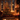 A warm and inviting indoor scene featuring a brightly lit Christmas tree decorated with red and gold ornaments, casting a festive glow. Numerous flickering candles are scattered on a polished wooden floor and an ornate white fireplace mantle, where wrapped gifts are also visible. Through a large window, a dark night sky with soft, bokeh-like snow is visible, enhancing the cozy and magical winter atmosphere.