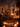 A warm and inviting indoor scene featuring a brightly lit Christmas tree decorated with red and gold ornaments, casting a festive glow. Numerous flickering candles are scattered on a polished wooden floor and an ornate white fireplace mantle, where wrapped gifts are also visible. Through a large window, a dark night sky with soft, bokeh-like snow is visible, enhancing the cozy and magical winter atmosphere.