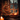 A warm and inviting indoor scene featuring a beautifully decorated Christmas tree positioned next to a wooden-framed window on a dark winter evening. The green fir tree is adorned with numerous glowing golden string lights and shiny red and gold spherical ornaments. Multiple lit pillar candles on the windowsill and in the foreground cast a soft, golden light, enhancing the cozy atmosphere, while outside the window, blurred city lights or other decorations create a bokeh effect against the cool night sky.