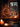A warm and inviting indoor scene featuring a beautifully decorated Christmas tree positioned next to a wooden-framed window on a dark winter evening. The green fir tree is adorned with numerous glowing golden string lights and shiny red and gold spherical ornaments. Multiple lit pillar candles on the windowsill and in the foreground cast a soft, golden light, enhancing the cozy atmosphere, while outside the window, blurred city lights or other decorations create a bokeh effect against the cool night sky.