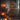 A warm and inviting interior scene featuring a fully decorated Christmas tree on the right and a glowing brick fireplace on the left. Numerous candles of varying sizes are lit on the mantelpiece, hearth, and floor, casting a soft, golden light throughout the room. Wrapped gifts are visible at the base of the Christmas tree, which is adorned with red baubles and tiny lights, creating a festive and cozy holiday atmosphere in a rustic setting.