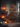 A warm and inviting interior scene featuring a fully decorated Christmas tree on the right and a glowing brick fireplace on the left. Numerous candles of varying sizes are lit on the mantelpiece, hearth, and floor, casting a soft, golden light throughout the room. Wrapped gifts are visible at the base of the Christmas tree, which is adorned with red baubles and tiny lights, creating a festive and cozy holiday atmosphere in a rustic setting.