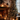 A cozy, warmly lit living room decorated for Christmas. A large, green Christmas tree, adorned with golden and silver ornaments and numerous twinkling lights, stands on the right next to a dark window. On the left, a rustic brick fireplace glows with a burning fire, flanked by stacked firewood. Multiple lanterns with lit candles and neatly wrapped brown paper gifts are arranged on the floor and a wooden mantelpiece, enhancing the festive and inviting holiday atmosphere.