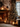 A cozy, warmly lit living room decorated for Christmas. A large, green Christmas tree, adorned with golden and silver ornaments and numerous twinkling lights, stands on the right next to a dark window. On the left, a rustic brick fireplace glows with a burning fire, flanked by stacked firewood. Multiple lanterns with lit candles and neatly wrapped brown paper gifts are arranged on the floor and a wooden mantelpiece, enhancing the festive and inviting holiday atmosphere.