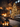 A vertical shot of a beautifully decorated rustic living room during Christmas. A grand stone fireplace with a roaring fire and garland-adorned mantelpiece is on the left, balancing a tall, heavily lit Christmas tree on the right, adorned with various ornaments. The room features exposed wooden beams, a comfortable sofa, and numerous glowing candles in lanterns and on surfaces, casting a warm, festive glow over the patterned rug and wooden floors.