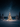 A beautifully illuminated Christmas tree covered in white snow stands centrally in a vast, pristine winter landscape under a dark, star-filled night sky. Warm golden lights adorn the evergreen tree, casting a gentle glow on the surrounding snow, while distant snow-covered forest trees line the horizon under the deep blue starry expanse.