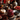 A close-up of a glass jar filled with bright red cranberry sauce, sitting on a dark wood coaster. The scene includes a small white pumpkin, bowls of cranberries and black currants, dried leaves, and a burning candle, all set on a dark wooden surface with a textured beige cloth. The backdrop features blurred flowers and a silver-colored decorative sphere, contributing to a warm and festive autumn ambiance.