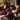 A warmly lit still life features a glass jar of cranberry sauce sitting on a small plate, surrounded by loose cranberries and sprigs of thyme. A silver spoon rests beside the jar. In the background, other autumnal elements such as pumpkins, pinecones, and vases filled with fall foliage create a festive Thanksgiving scene on a rustic wooden surface.