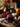A warmly lit still life features a glass jar of cranberry sauce sitting on a small plate, surrounded by loose cranberries and sprigs of thyme. A silver spoon rests beside the jar. In the background, other autumnal elements such as pumpkins, pinecones, and vases filled with fall foliage create a festive Thanksgiving scene on a rustic wooden surface.