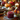 A close-up shot of cranberry sauce in a glass jar, placed on a wicker placemat with a blue-gray cloth napkin, as part of a Thanksgiving table setting. The background contains various autumnal decorations, including pumpkins, pinecones, flowers, and a lit candle, adding a warm glow. A silver spoon rests in the cranberry sauce.