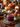 A close-up shot of cranberry sauce in a glass jar, placed on a wicker placemat with a blue-gray cloth napkin, as part of a Thanksgiving table setting. The background contains various autumnal decorations, including pumpkins, pinecones, flowers, and a lit candle, adding a warm glow. A silver spoon rests in the cranberry sauce.