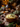 A rustic bowl of mashed potatoes topped with rosemary sits on a burlap cloth surrounded by autumn decorations. Miniature pumpkins, acorns, pinecones, and fall-colored flowers are arranged on a wooden table. The image evokes a warm, cozy, and inviting atmosphere perfect for Thanksgiving or fall-themed content.