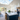 A wide-angle interior shot of a modern, open-concept kitchen and living room. A mother holding a toddler stands next to the father on the right, looking towards a large navy blue kitchen island with a white marble top. The space is brightly illuminated by a large vaulted skylight, recessed lighting, and natural light from sliding glass doors leading to a terraced backyard with lush greenery and colorful autumn foliage.
