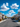 A bright, sunny view down a tree-lined suburban street featuring modern, light-colored two-story houses with garages on both sides. The asphalt road is flanked by sidewalks and numerous parked cars, all under a clear blue sky dotted with fluffy white clouds. The scene conveys a peaceful and well-established residential neighborhood with a focus on architecture and daily life.