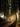 A silhouetted woman with a ponytail runs down a paved road, followed closely by a medium-sized dog, during an early morning or late afternoon with golden light. Tall, leafy trees line both sides of the road, forming an avenue, and strong sunbeams cut through a visible mist or haze, illuminating the scene from the right side. Long, dark shadows stretch across the road, emphasizing the low angle of the sun and the peaceful, active mood of the rural setting.